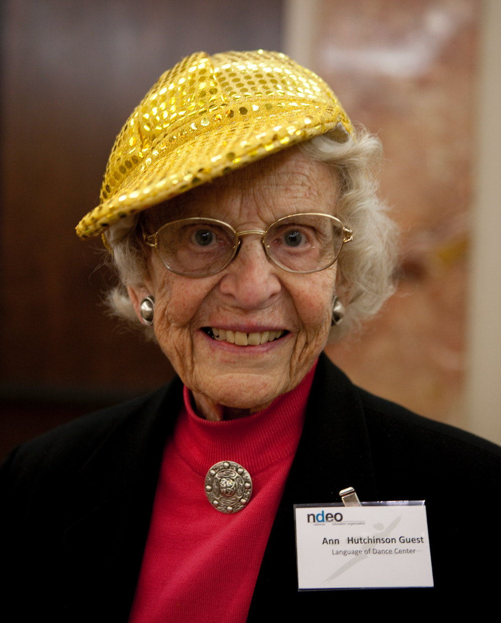 Ann wears her signature sparkly gold cap, glasses, a red turtleneck with a broach, and a black jacket and smiles at the camera.