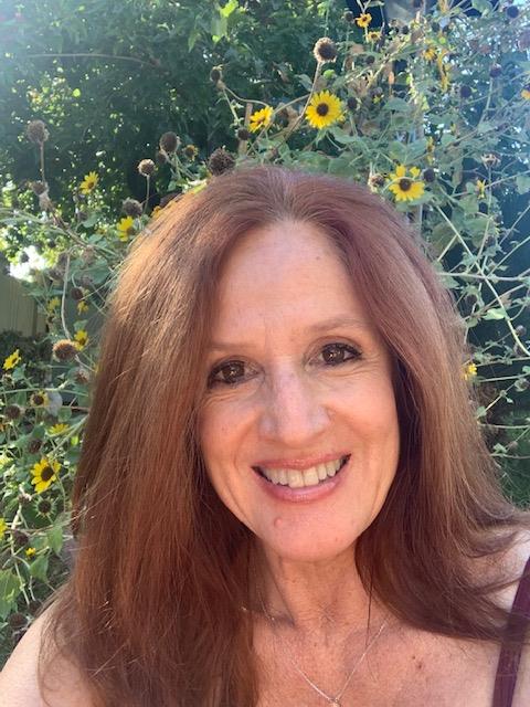 MaryAnne has shoulder-length brown hair, is smiling, and is pictured before flowers on a sunny day.