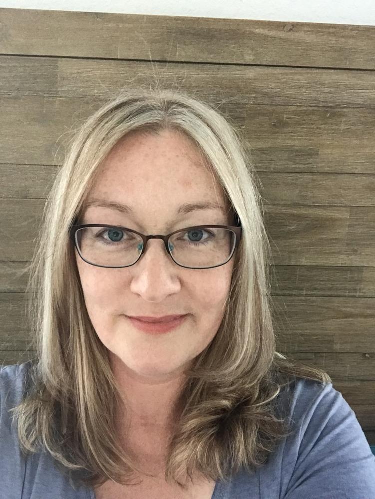 Dawn is a white woman with shoulder length light hair, wearing glasses, a blue shirt, and is pictured before a wooden background.