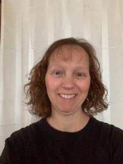 She is a white woman with short curly hair, smiling and wearing a dark shirt.