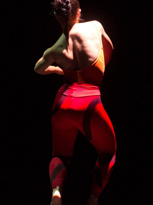 A dancer in red leggings and a yellow leotard, facing away from the camera, stands against a black backround.