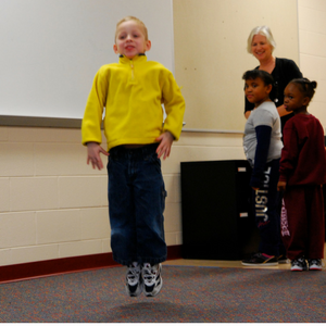 A young boy in a yellow long sleeve shirt jumps up in the air while two young students and a teacher watch.