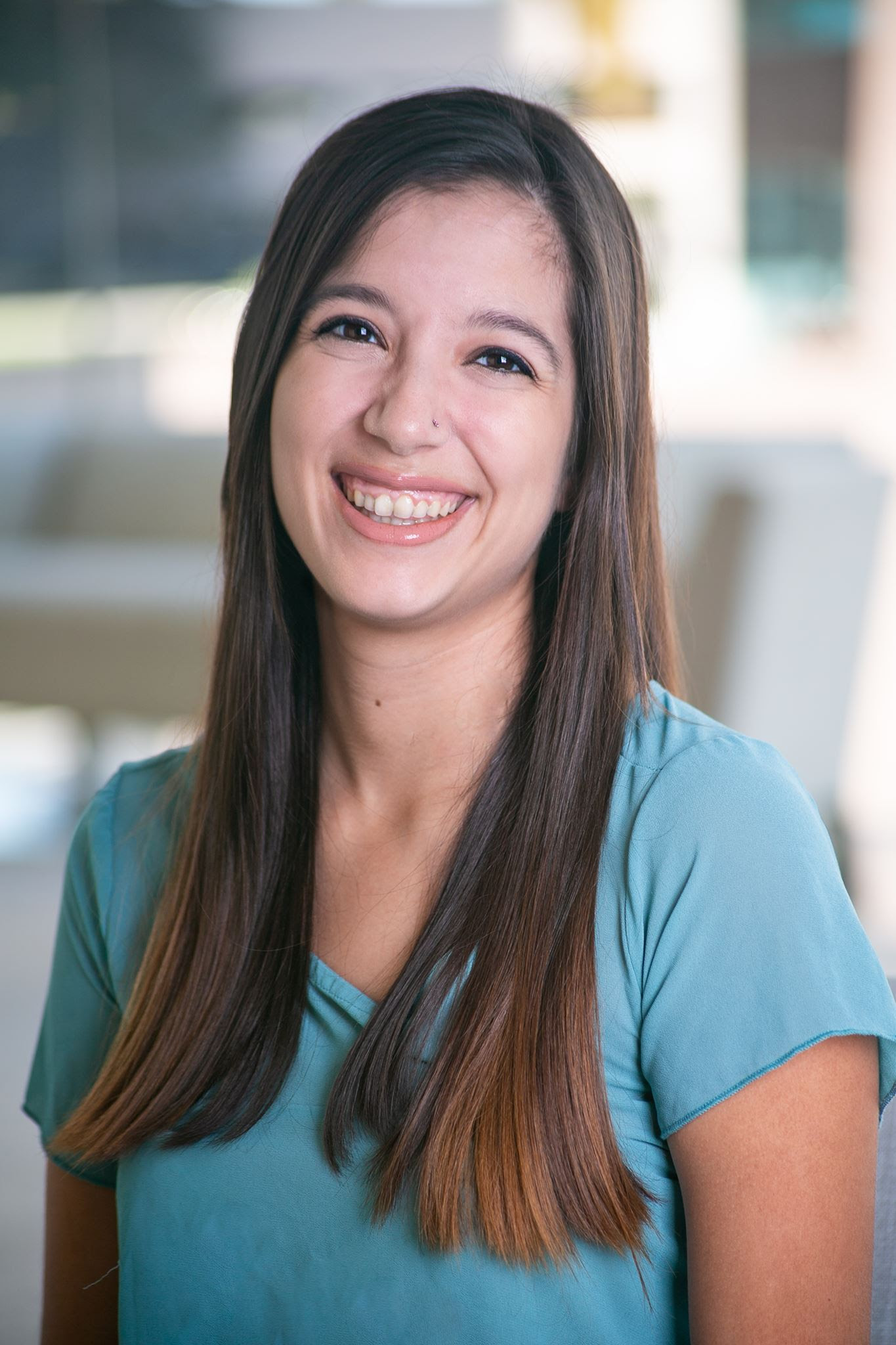 A head shot of Caitlin, a woman with brown hair, smiling with a blue shirt on against a blurry back round.