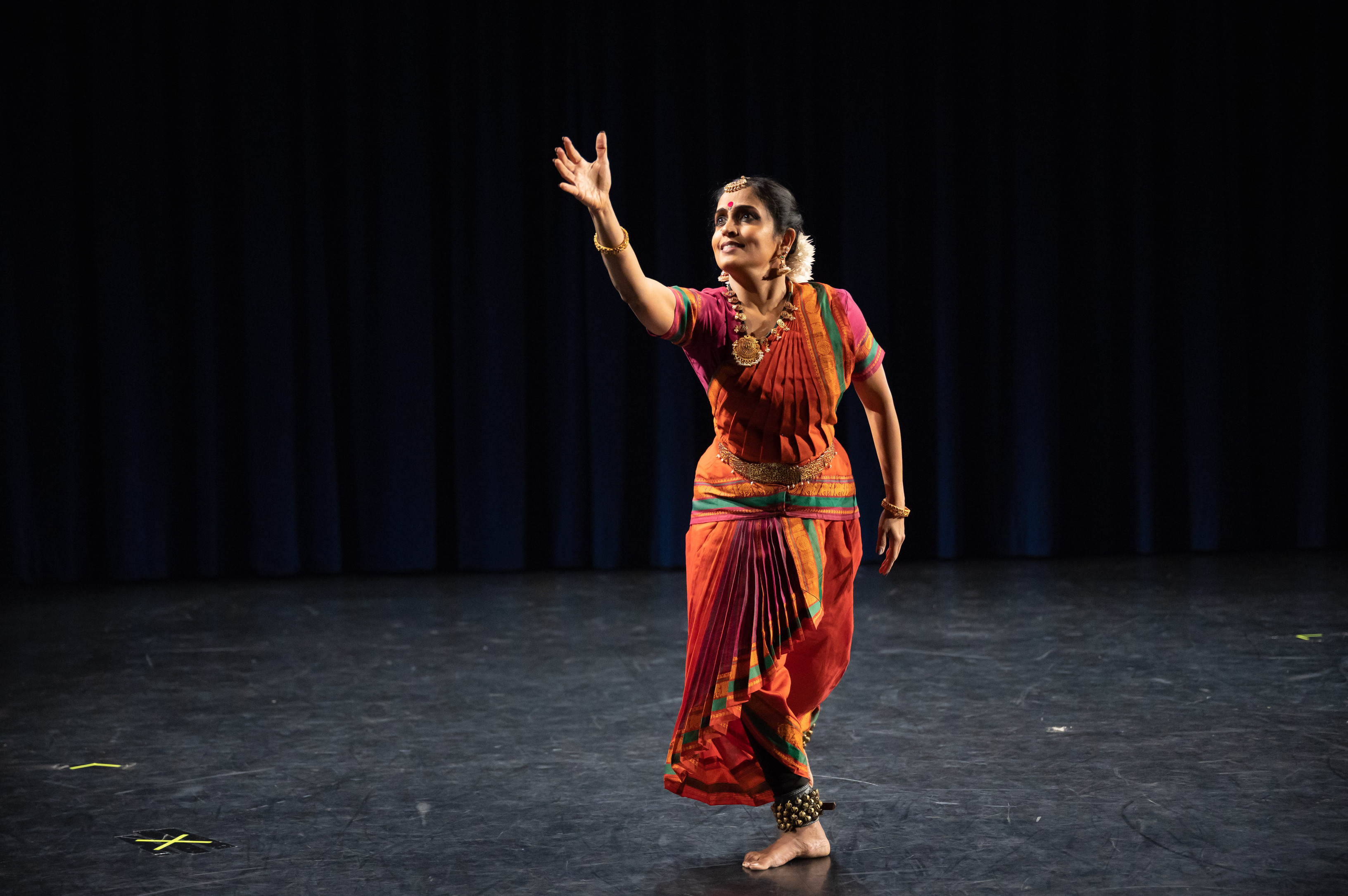 A photo of an Indian woman on stage in a pink and oranige dress, with a large necklace and earrings, her right arm is outreached.