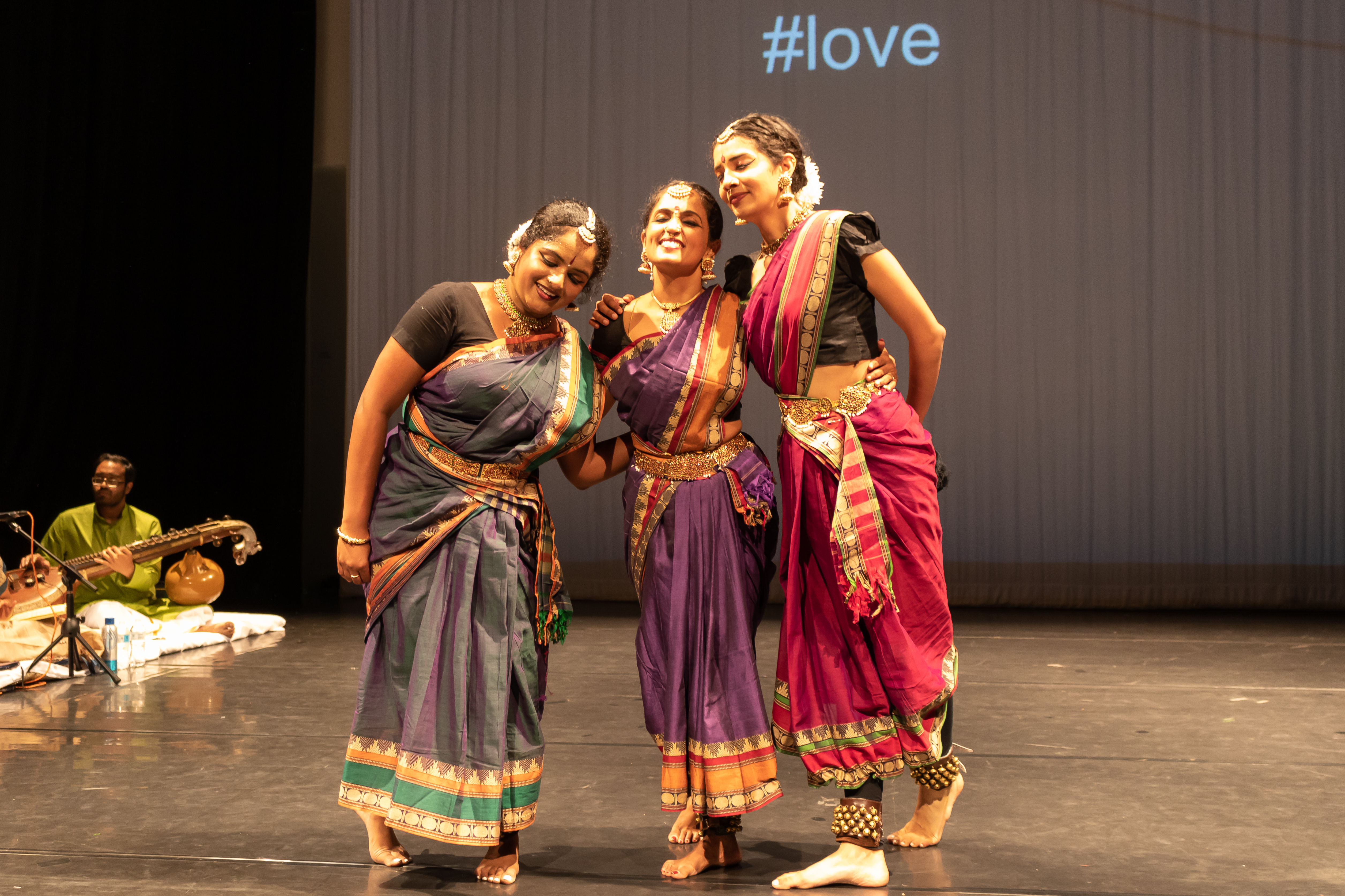 Three Indian women in colorful dresses leaning in towards each other smiling.