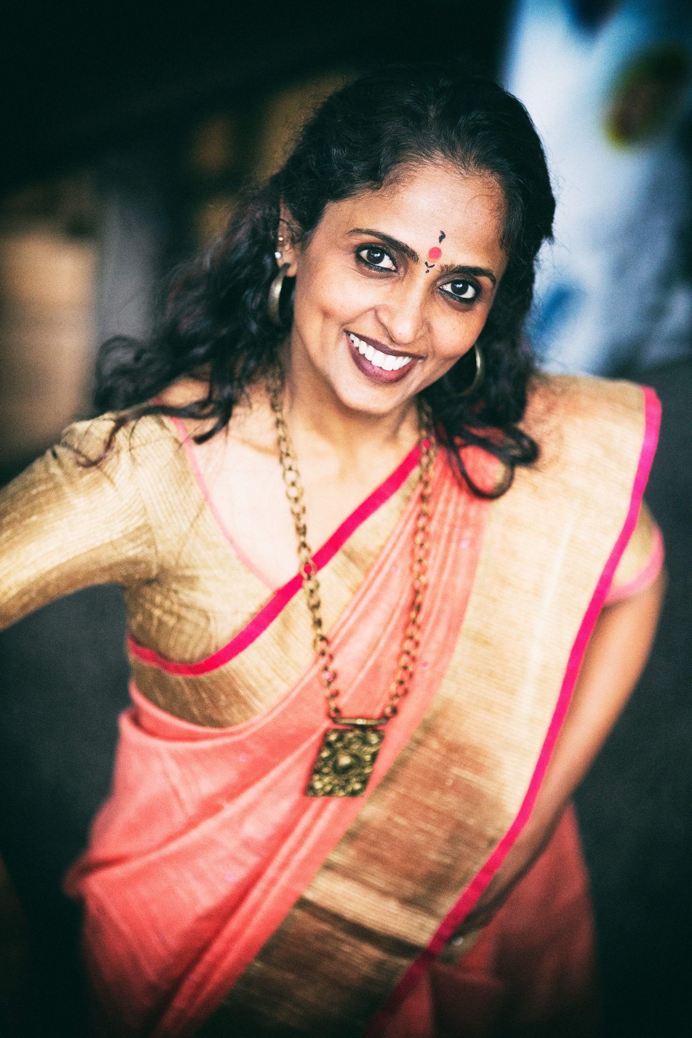 A photo of Deepa, she has dark brown hair, is in orange clothing, and against a blurred back round.