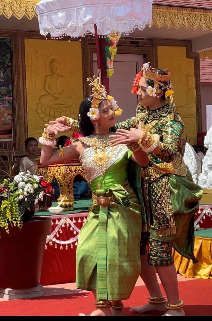 Two dancers dressed in colorful clothing and head dresses are standing closley posing, and looking at each other smiling.