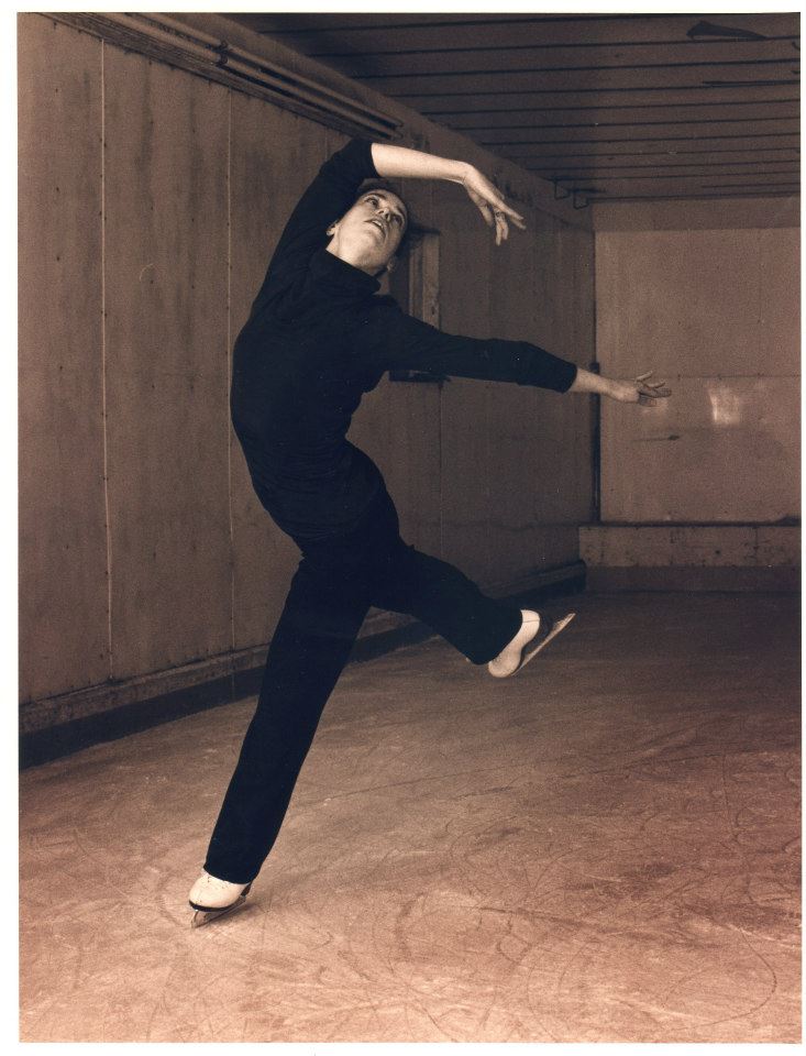 A black and white photo of Heather, wearing all black, on figure skates, posing with one arm over head.