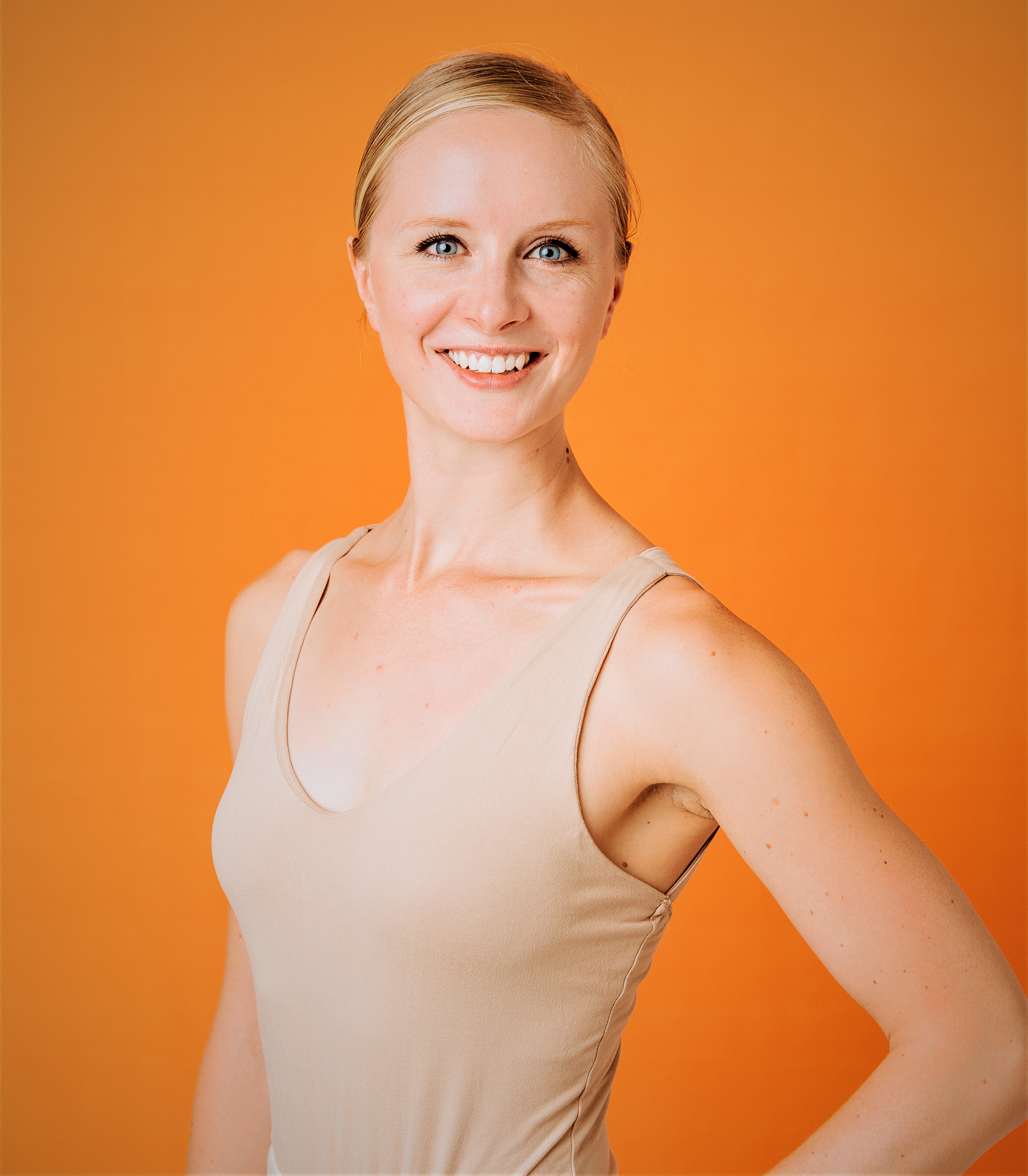 A head shot of Krista, a white woman with blonde hair. She is wearing a light brown tank top, and is standing with one hand on her hip against an organge backround.