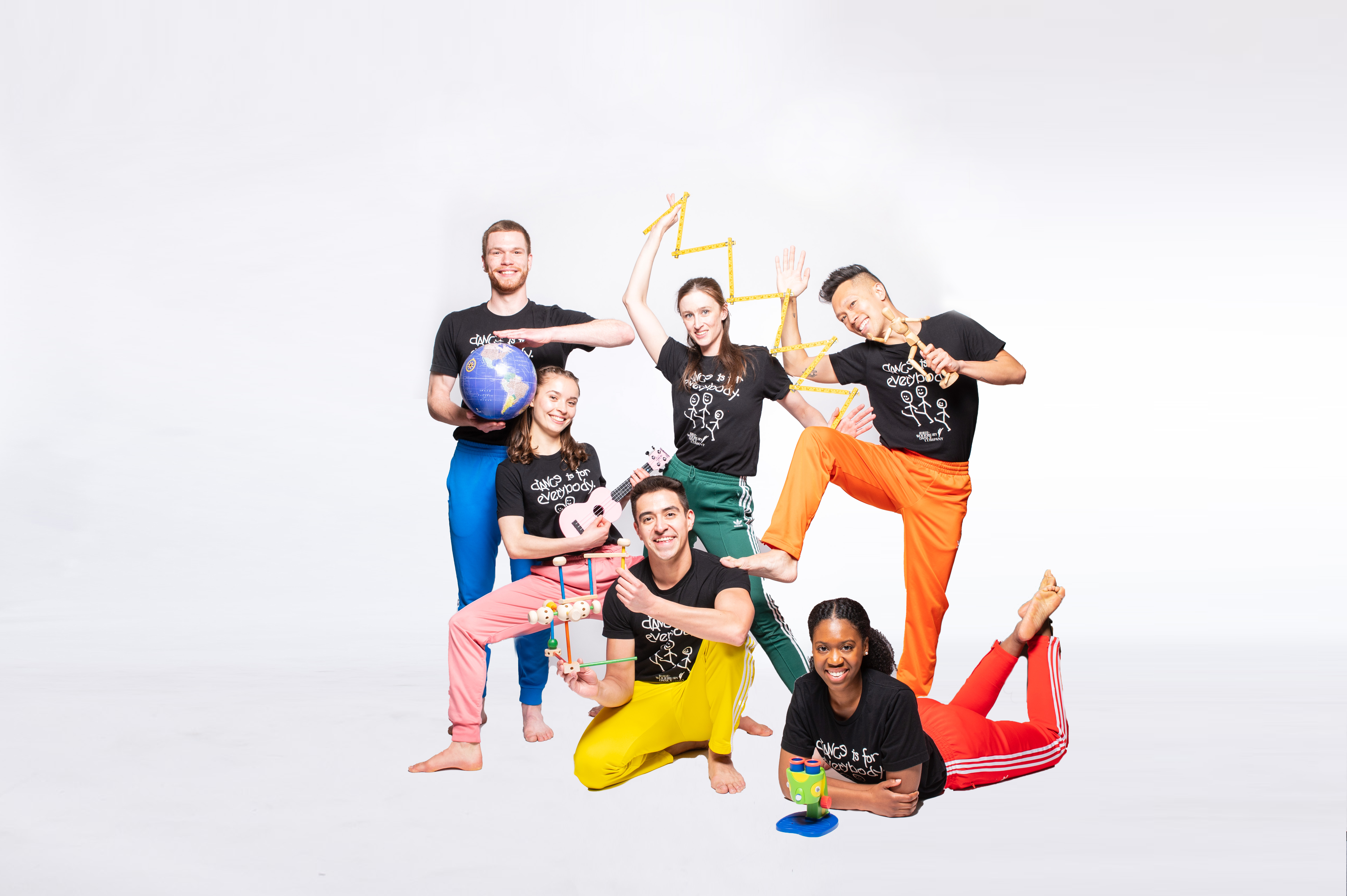 A group of adult dancers in colorful pants and black tshirts, holding various objects and posing against a white backround