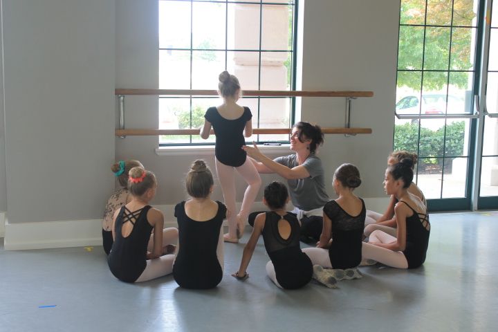 A small group of young ballet dancers circles around another dancer demonstrating a step with a teacher helping her at the barre.