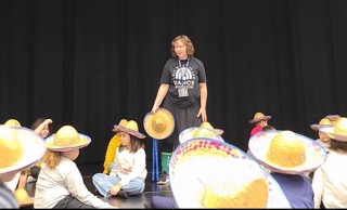 Photo of Gina standing in front of a group of kids with large hats on. She is wearing all black and standing aginst a black curtain.