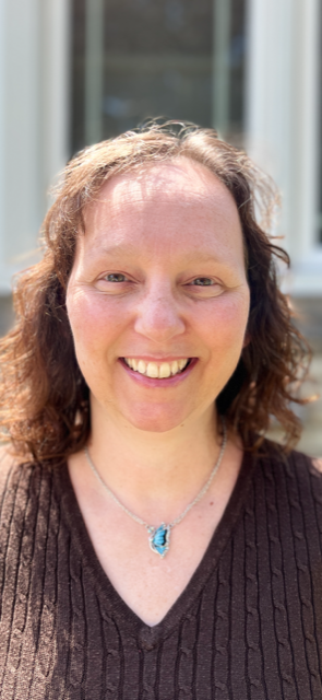 headshot of Gina, a white woman with light brown hair. She is smiling at the camera, is wearing a silver necklace and brown shirt.
