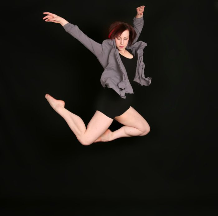 Helen jumping with her legs tucked back and her arms above her head. She is wearing a black biketard and gray sweater against a black back round.