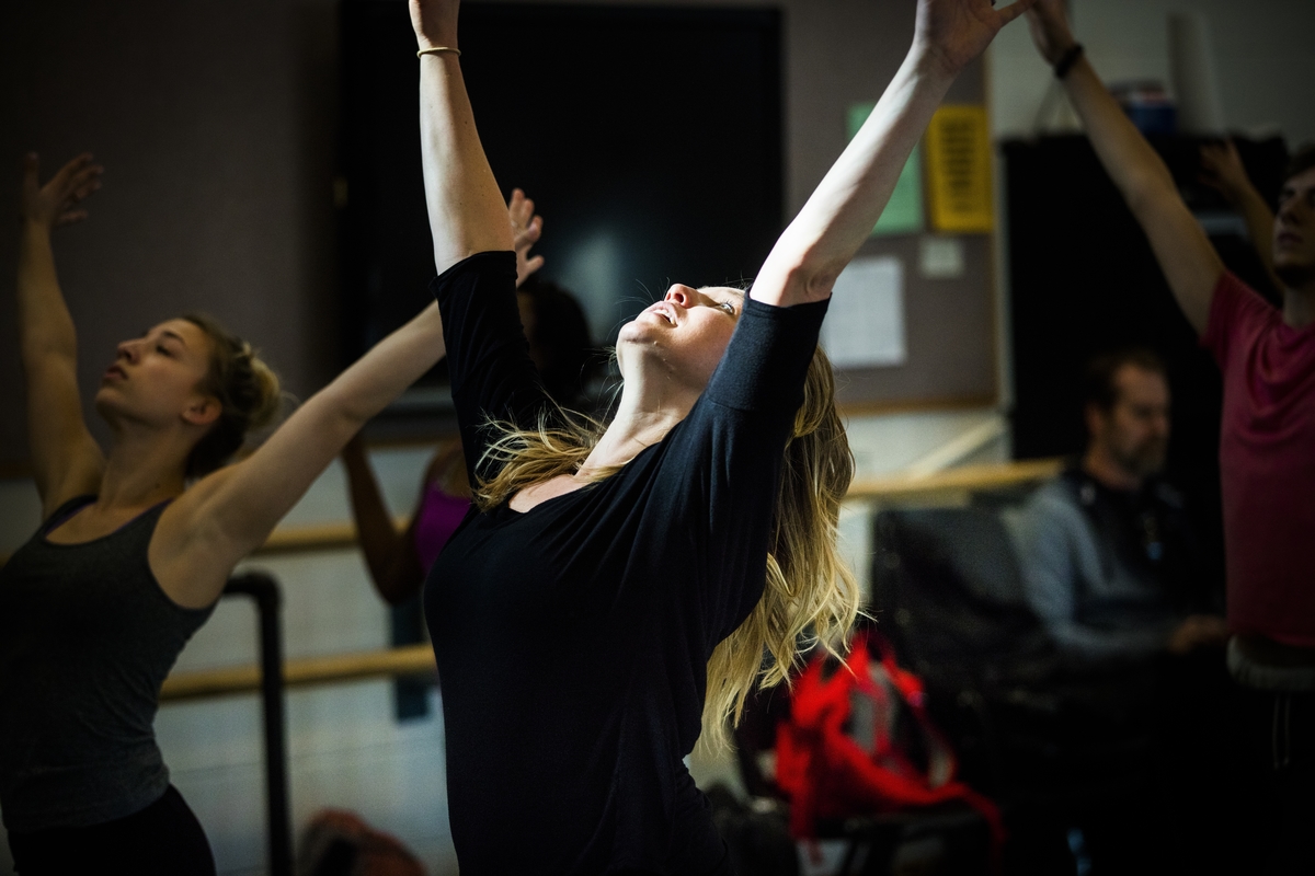 a white woman with blonde hair, wearing a black tshirt, she has both arms up in a v shape and is looking at the ceiling