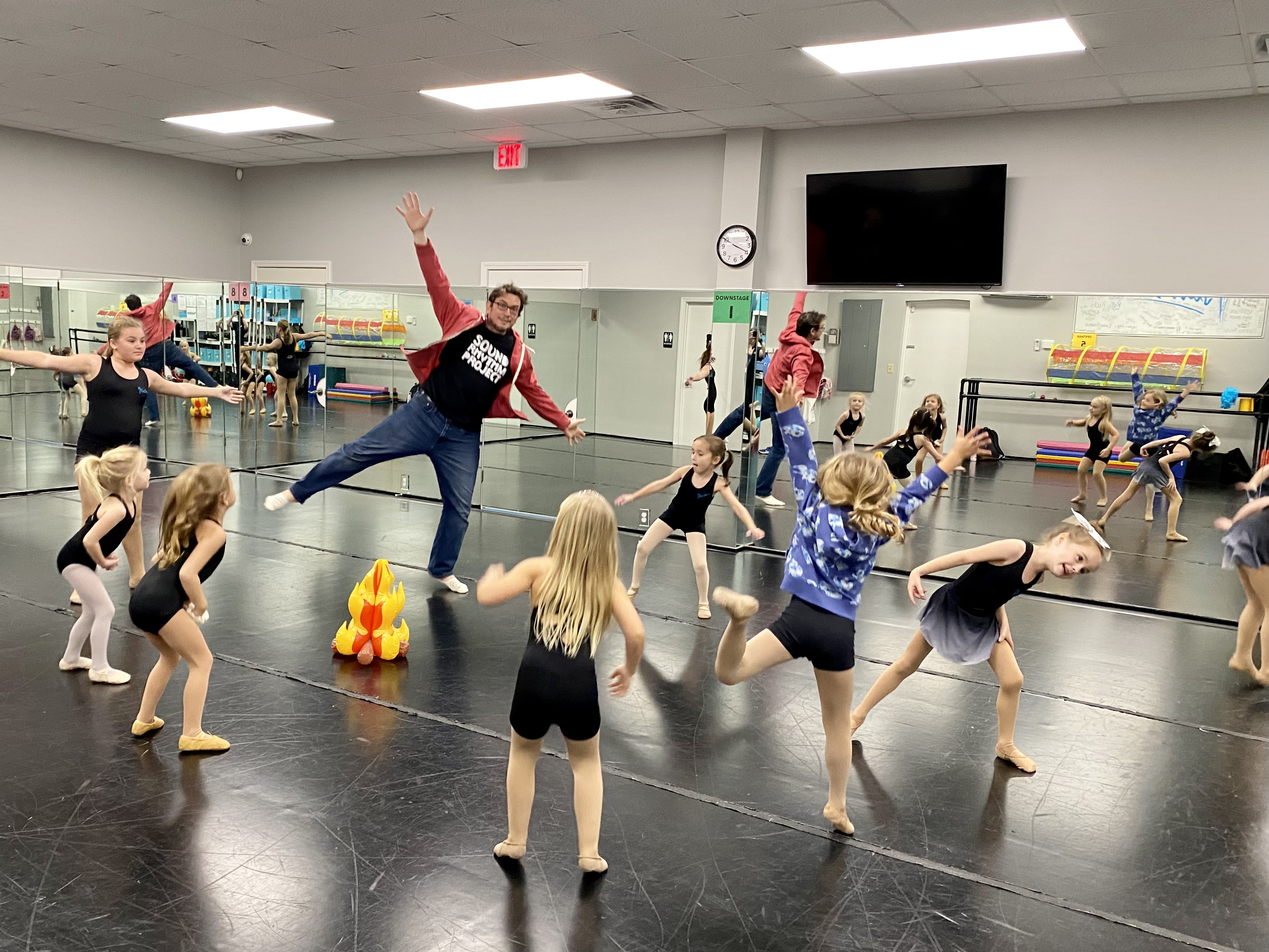 a teacher leads a class of very young dancers, they are all in various shapes and wearing dance clothes