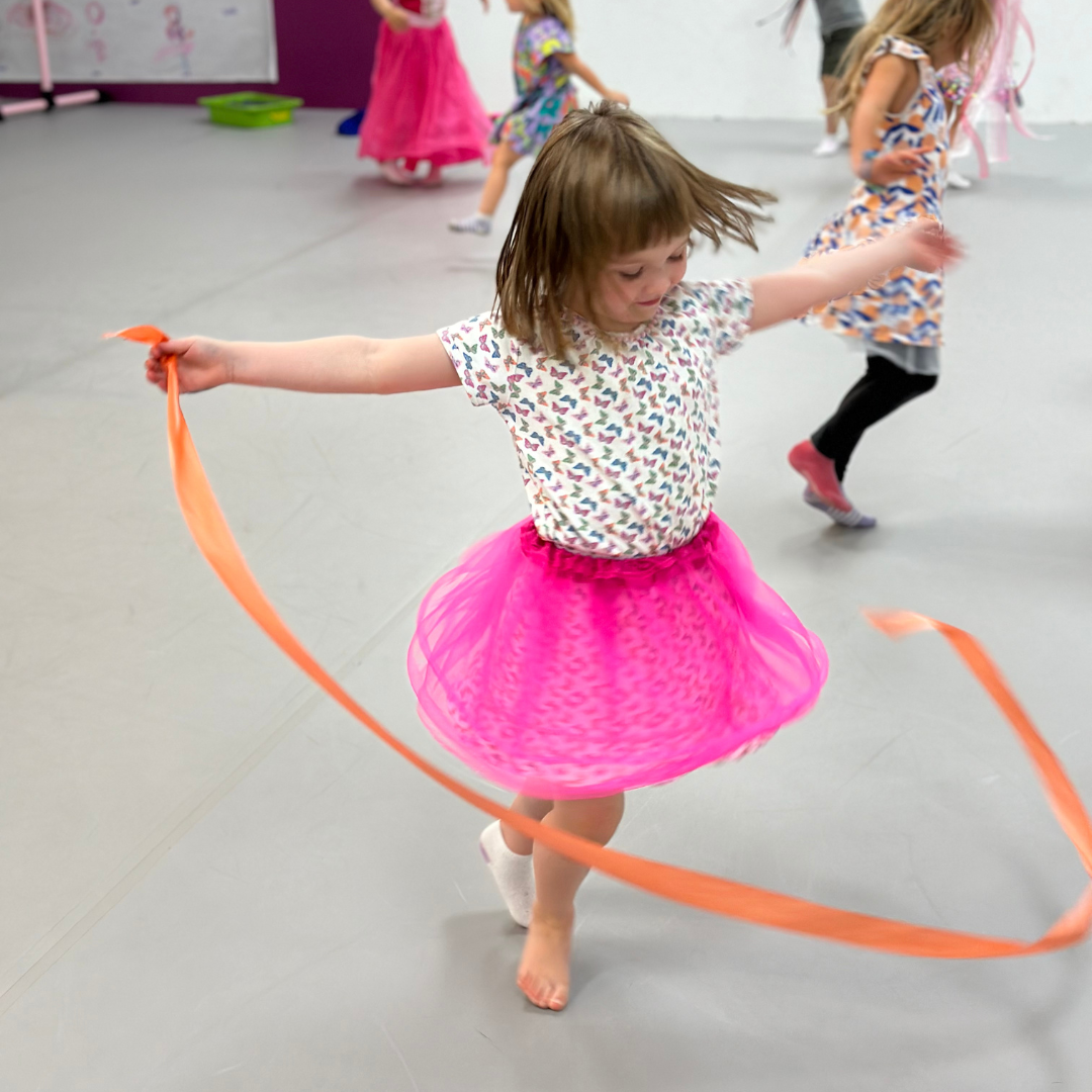 A young dancer twirls with an orange ribbon in class with a bright pink tutu, a flower patterned shirt with other students moving in the backround.