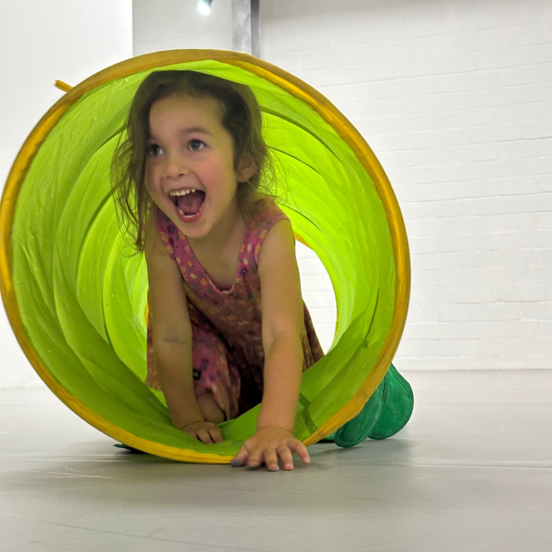 A young student with light colored hair and a pink dress crawling through a bright green tube with a smile on her face.