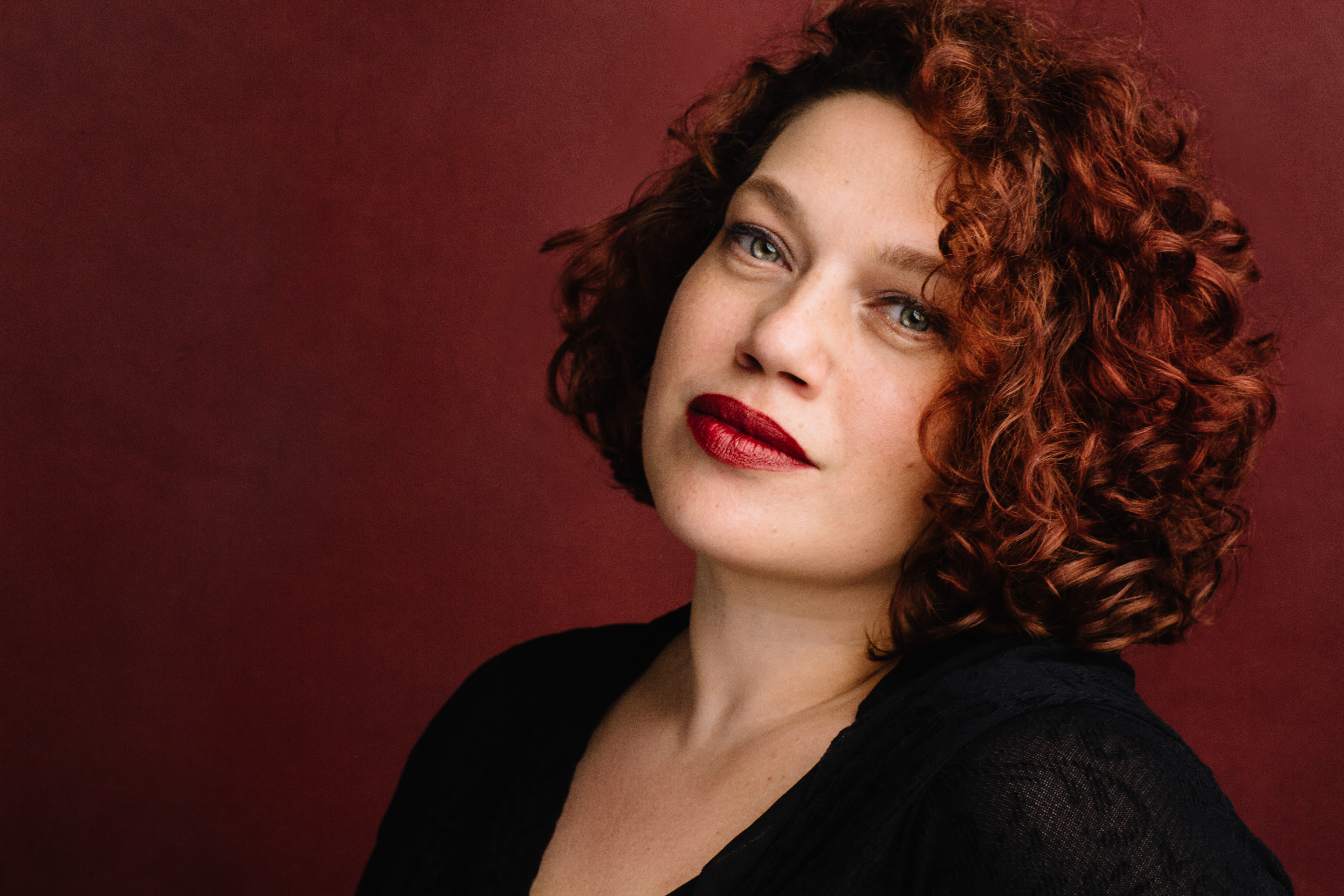 Headshot of Alexandra, a white woman with short red curly hair. She has on red lipstick, head slightly tilted back staring into the camera. She is wearing a black top and the back round is burgundy.
