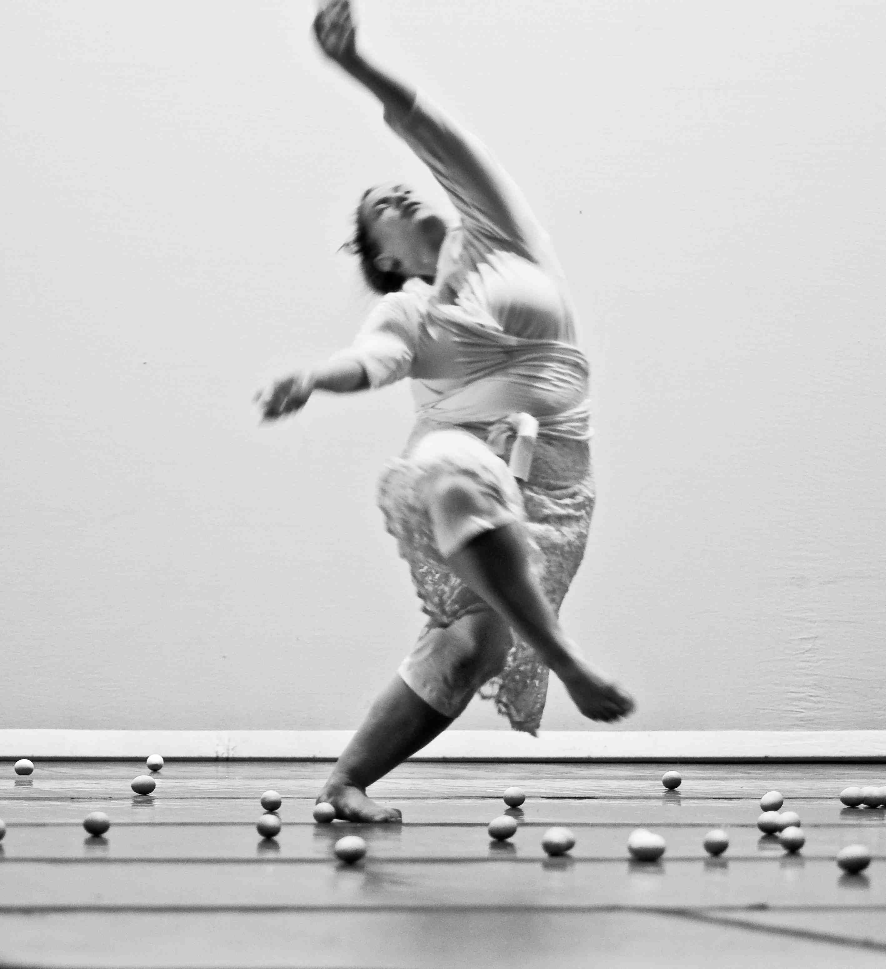 Black and white photo of Alexandra dancing.  She is in a wrap top and skirt, standing on one leg, the other leg is lifted and crossing over her standing leg. Her arms are lifted above her.
