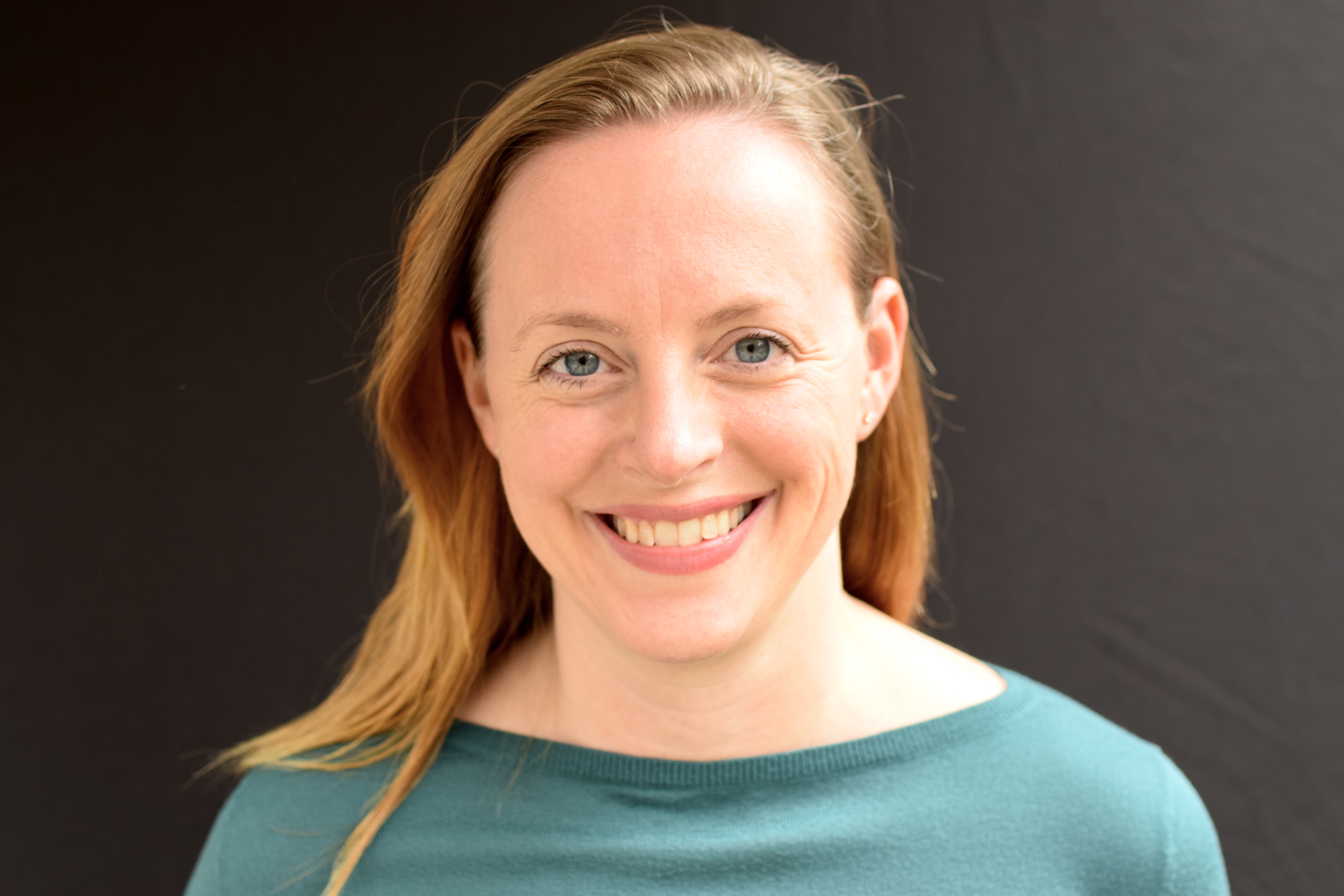 Photo of Ashley, a white woman with blondish hair. She is in front of a black backround, wearing a teal shirt, smiling directly into the camera.