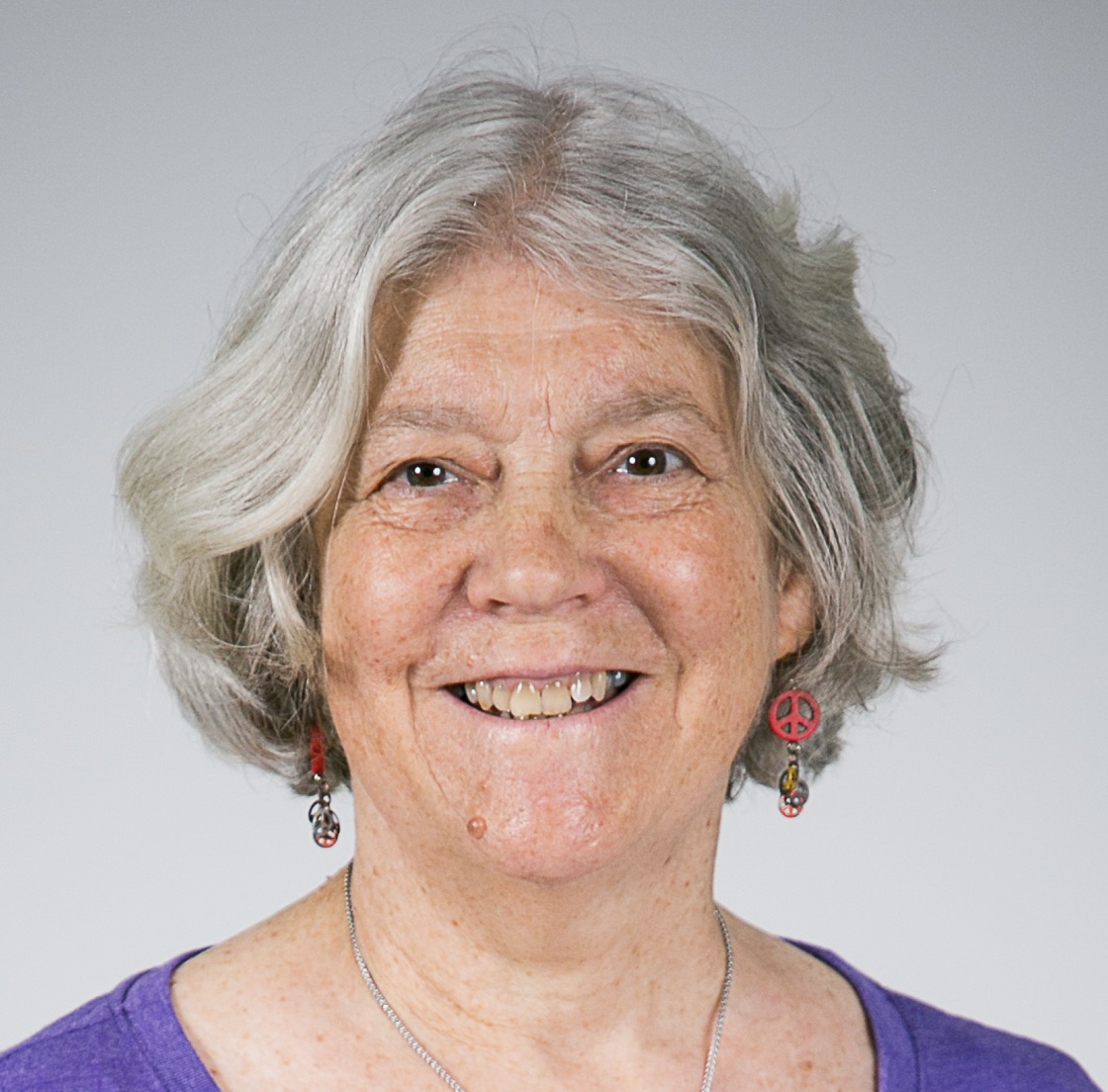 Sandi has chin length gray hair, a big smile, and is wearing peace sign earrings and a purple shirt